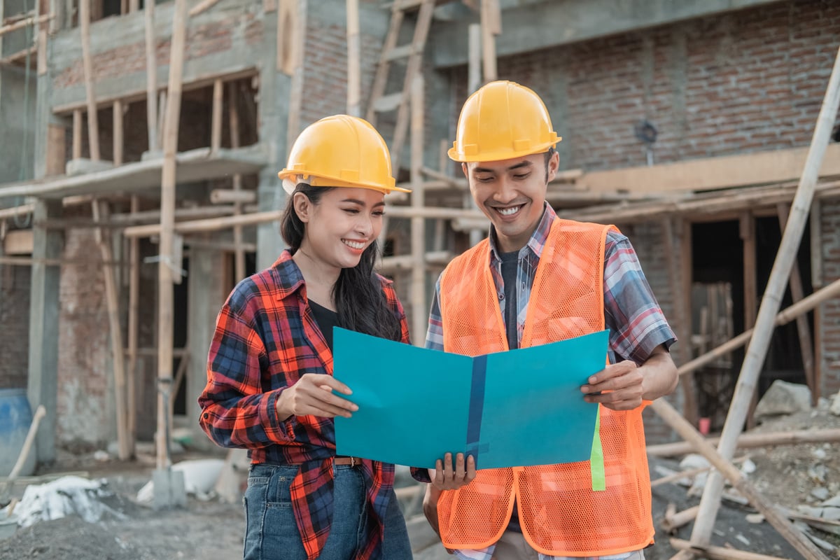 Male and Female Contractors Standing Holding a Smiling Sketch Book Wearing a Safety Helmet